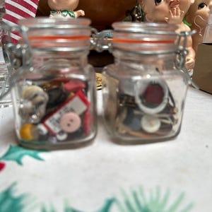 May include: Two small, clear glass jars with metal clasps and orange rubber seals, filled with assorted buttons and small objects. The jars sit on a patterned tablecloth, with figurines in the background.