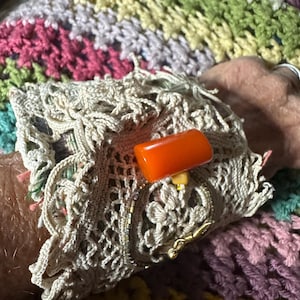 May include: A beige crocheted cuff bracelet with an orange rectangular stone and gold-colored accents. The bracelet is worn on a wrist, set against a colorful crocheted blanket.