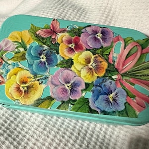 May include: A rectangular, turquoise-colored trinket box decorated with a floral design. The design features a bouquet of colorful pansies in shades of yellow, purple, and blue, accented with a pink bow and a butterfly.