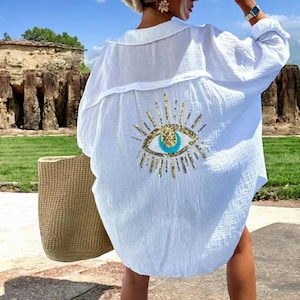 Op de afbeelding: Witte, oversized blouse met een gouden en turquoise oogdesign op de rug. De blouse heeft lange mouwen en een kraag. Een geweven draagtas is zichtbaar aan de linkerkant. Gemaakt van een lichte stof, ideaal voor de zomer.