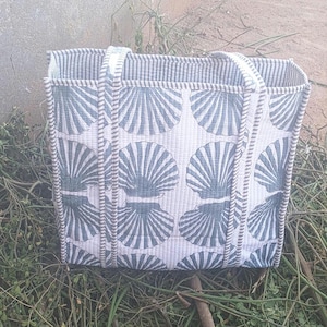 Könnte beinhalten: Eine rechteckige Tragetasche mit weißem Hintergrund und einem sich wiederholenden Muster aus blauen Muscheldesigns. Die Tasche hat zwei Griffe und ist mit einem gestreiften Stoff verziert. Die Tasche liegt auf grünem Laub.