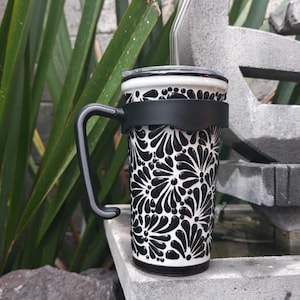 Puede incluir: Taza de viaje con estampado floral en blanco y negro, asa y tapa negras. La taza tiene una banda negra y una pajita de cristal transparente. Está sobre una superficie de piedra gris.