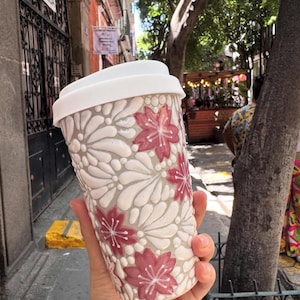 May include: A white and gray ceramic travel mug with a white lid. The mug features a raised floral design with red flowers. The mug is held in a hand, with a blurred outdoor background.