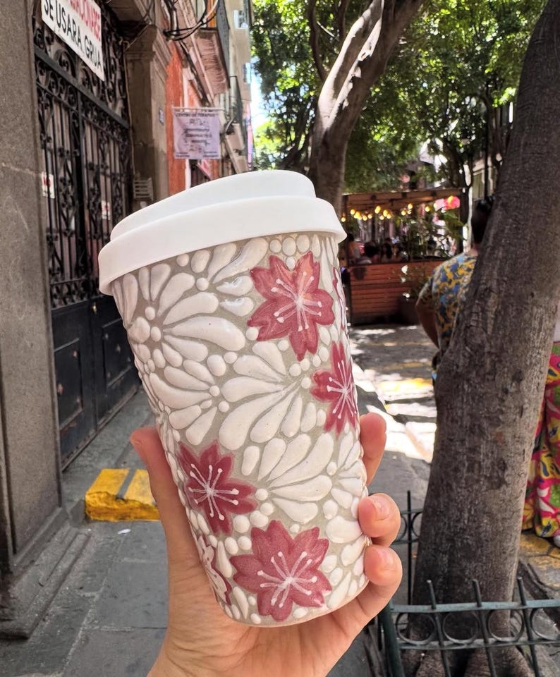 May include: A white and gray ceramic travel mug with a white lid. The mug features a raised floral design with red flowers. The mug is held in a hand, with a blurred outdoor background.