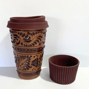 May include: A brown ceramic travel mug with a decorative floral pattern and a matching brown lid. A separate brown silicone sleeve sits next to the mug. The mug is designed to hold hot or cold beverages.
