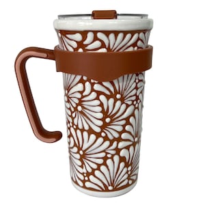 May include: A ceramic travel mug with a brown and white floral design. The mug has a brown handle and a brown silicone band around the middle. The lid is clear with a brown slider.