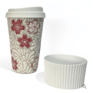 May include: A ceramic travel mug with a white lid and a matching sleeve. The mug is decorated with a floral pattern in red and white on a gray background. The sleeve is white with vertical ridges.