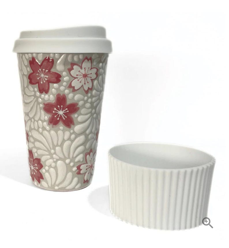 May include: A ceramic travel mug with a white lid and a matching sleeve. The mug is decorated with a floral pattern in red and white on a gray background. The sleeve is white with vertical ridges.
