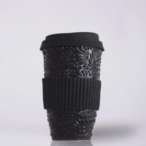 May include: Black travel mug with a floral embossed design. The mug has a black silicone sleeve and a black lid. The mug is designed to keep beverages warm or cold. The mug is made of ceramic.
