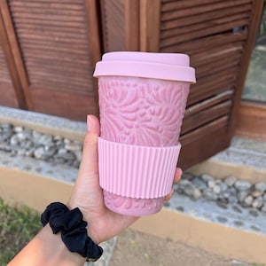 May include: A pink travel mug with a floral design, a matching lid, and a ribbed sleeve. The mug is held in a hand, with a black scrunchie on the wrist. The background shows a wooden door.