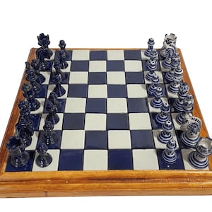 Puede incluir: Un juego de ajedrez completo con marco de madera. El tablero presenta baldosas azules y blancas. Las piezas de ajedrez son azul oscuro y blanco, con diseños detallados. El juego está listo para jugar.
