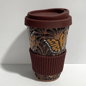 May include: A brown travel mug with a lid and a ribbed sleeve. The mug is decorated with orange and white butterfly designs on a dark brown background. The lid and sleeve are a solid brown color.