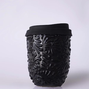 Puede incluir: Un termo negro con un diseño floral en relieve. El termo tiene una tapa negra y una superficie texturizada. El termo está diseñado para mantener las bebidas calientes o frías. El termo es de cerámica.