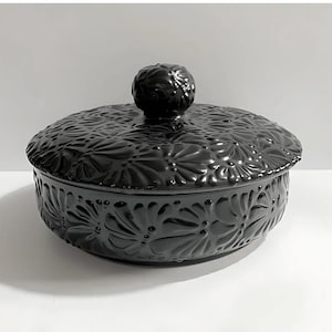 May include: A black ceramic lidded bowl with a floral embossed design. The round bowl has a matching lid with a spherical knob. The bowl and lid have a glossy finish and are set against a white background.