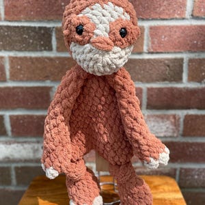 May include: A handmade, plush sloth toy in shades of coral, cream, and black. The sloth has a textured, chunky knit appearance, with a cream-colored face and paws, and black button eyes. The toy is posed on a wooden surface against a brick background.