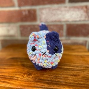 May include: A handmade, crocheted cat figurine. The cat is primarily light blue with colorful flecks, and a dark blue section on its head. It has black safety eyes and a stitched mouth. The cat is sitting on a wooden surface.