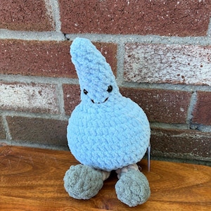 May include: A light blue, teardrop-shaped plush toy made of soft, textured yarn. It features two small black bead eyes and a stitched black mouth. The plush has two rounded, grey-coloured feet extending from the bottom of its round body.
