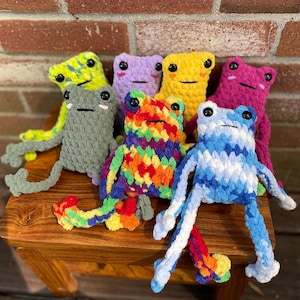 May include: A collection of handmade crochet frog plushies in various colors, including green, purple, yellow, pink, blue, and rainbow. Each frog has black button eyes and is posed on a wooden surface.
