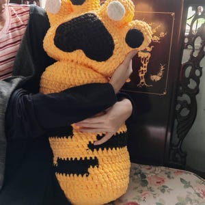 Puede incluir: Una almohada grande y afelpada con forma de pata, de ganchillo amarillo y negro. La almohada presenta un cuerpo amarillo con detalles negros y almohadillas blancas. El artículo está diseñado para ser abrazado.