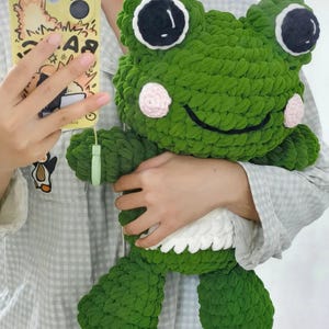 May include: A large, plush green frog toy with big black and white eyes, pink cheeks, and a smiling mouth. The frog is made of a soft, textured yarn and has white detailing on its belly. A cute, handmade toy.