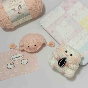 Puede incluir: Una colecci&oacute;n de art&iacute;culos de ganchillo hechos a mano, que incluyen un cerdo rosa con detalles florales, un h&aacute;mster blanco sosteniendo una semilla de girasol y una tarjeta rosa de Hello Kitty con el texto "I missssss you". Tambi&eacute;n hay un ovillo de lana rosa y una tela estampada.