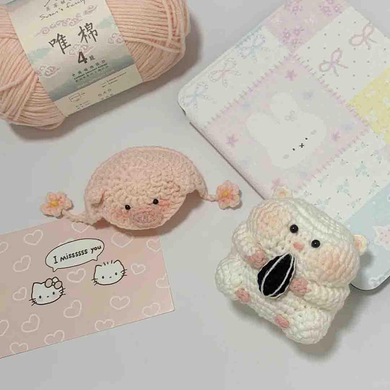 Puede incluir: Una colecci&oacute;n de art&iacute;culos de ganchillo hechos a mano, que incluyen un cerdo rosa con detalles florales, un h&aacute;mster blanco sosteniendo una semilla de girasol y una tarjeta rosa de Hello Kitty con el texto "I missssss you". Tambi&eacute;n hay un ovillo de lana rosa y una tela estampada.