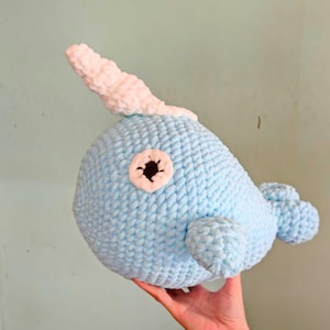 Może przedstawiać: Jasnoniebieska, pluszowa zabawka narwala z białym rogiem i detalami oczu. Ręcznie robiona pluszowa zabawka ma zaokrąglony korpus, małe płetwy i teksturowany, dziergany wygląd. Kapryśna i przytulna zabawka.