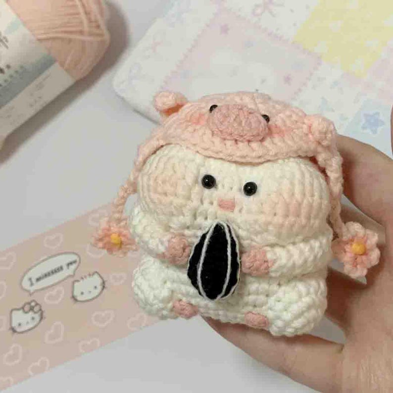 Puede incluir: Un h&aacute;mster de ganchillo hecho a mano con un gorro de cerdo rosa, sosteniendo una semilla de girasol negra y blanca. El h&aacute;mster es principalmente blanco con acentos rosas. El gorro tiene un hocico de cerdo y detalles de flores. El dise&ntilde;o es lindo y caprichoso.