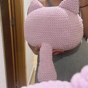 May include: A light pink, handmade cat-shaped pillow. The pillow features a round head with pointed ears and a long, cylindrical tail. The soft, plush material is a solid light pink color. The pillow is designed to be a decorative item or a cuddly toy.