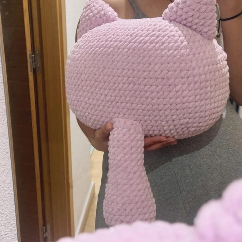 May include: A light pink, handmade cat-shaped pillow. The pillow features a round head with pointed ears and a long, cylindrical tail. The soft, plush material is a solid light pink color. The pillow is designed to be a decorative item or a cuddly toy.