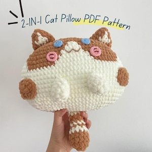 Könnte beinhalten: Ein gehäkeltes, katzenförmiges Kissen aus weißem und braunem Garn mit rosa kreisförmigen Wangen, blauen kreisförmigen Augen und einem kleinen braunen Schwanz. Der Text 2-IN-1 Cat Pillow PDF Pattern steht in blauer Schrift über dem Plüschobjekt.