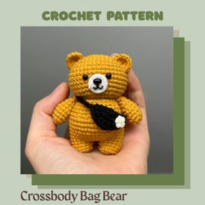 May include: A crocheted golden-yellow bear with a black crossbody bag and a white flower accent. The bear has black eyes and a black nose. The text "CROCHET PATTERN" and "Crossbody Bag Bear" are displayed.