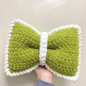 May include: A handmade, green bow-shaped pillow with a white border and center band. The pillow is made of a soft, textured material, and is held by a person wearing a blue long-sleeve shirt.