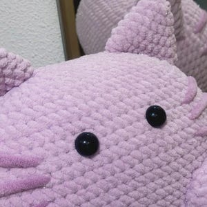 May include: Close-up of a handmade, lavender-colored crochet stuffed animal with black button eyes. The plush toy has textured, fluffy yarn and decorative accents. The image shows the head and part of the body of the toy.