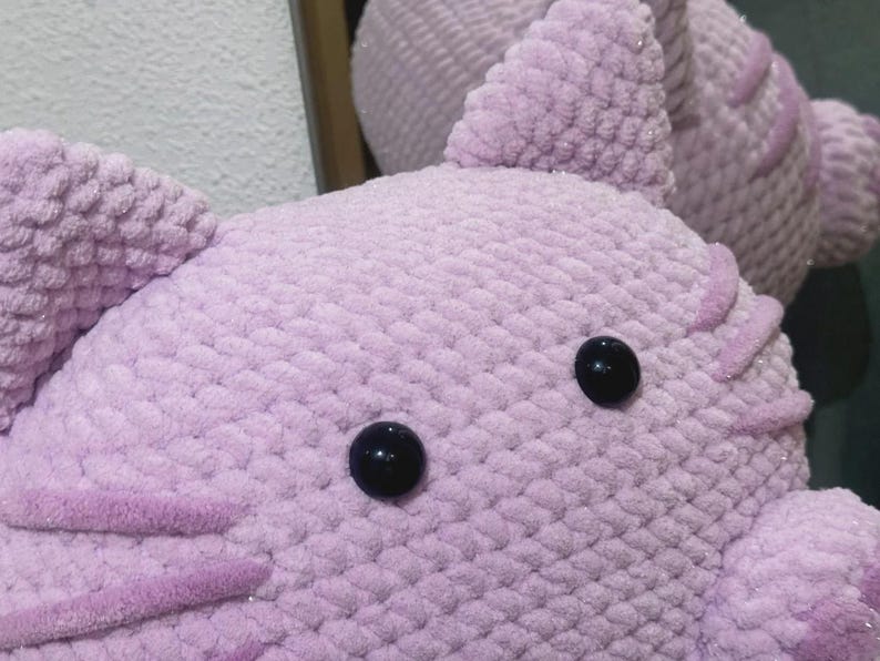 May include: Close-up of a handmade, lavender-colored crochet stuffed animal with black button eyes. The plush toy has textured, fluffy yarn and decorative accents. The image shows the head and part of the body of the toy.