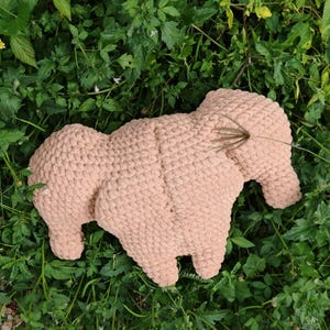 May include: A light brown, handmade, crocheted elephant-shaped pillow. The pillow is lying on a bed of green grass and foliage. The elephant has a textured, chunky knit appearance, with defined legs, ears, and a trunk.