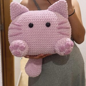 May include: A plush, light pink cat-shaped pillow with black button eyes and embroidered details. The pillow features a rounded head with pointed ears, pink stripes, and paw-shaped feet. The soft, cuddly design is perfect for a child's room or as a decorative item.
