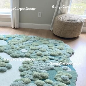 Puede incluir: Una alfombra inusual, de forma irregular, con un dise&ntilde;o texturizado en tres dimensiones. La alfombra presenta una mezcla de tonos verdes claros y oscuros, y azules, que recuerdan a un paisaje. Un otom&aacute;n redondo y tejido est&aacute; cerca. El texto "GangaCarpetDecor" es visible.