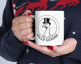 Könnte beinhalten: Weiße Keramiktasse mit einer Schwarz-Weiß-Illustration einer Gans mit Zylinder, umgeben von den Worten "SILLY GOOSE". Die Tasse wird von einer Person gehalten, die einen marineblauen Pullover mit einem roten Rentier-Muster trägt.