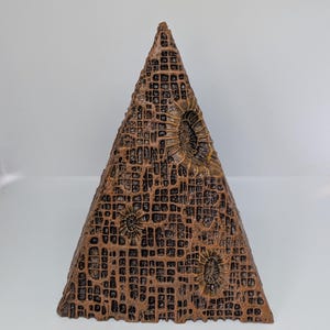 K&ouml;nnte beinhalten: Eine bronzefarbene, pyramidenf&ouml;rmige Skulptur mit einer strukturierten, gitterartigen Oberfl&auml;che. Die Pyramide weist kreisf&ouml;rmige, fossilartige Muster auf und ist vor einem wei&szlig;en Hintergrund platziert. Das Design deutet auf ein einzigartiges Dekorationsst&uuml;ck hin.