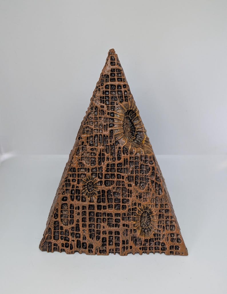 K&ouml;nnte beinhalten: Eine bronzefarbene, pyramidenf&ouml;rmige Skulptur mit einer strukturierten, gitterartigen Oberfl&auml;che. Die Pyramide weist kreisf&ouml;rmige, fossilartige Muster auf und ist vor einem wei&szlig;en Hintergrund platziert. Das Design deutet auf ein einzigartiges Dekorationsst&uuml;ck hin.