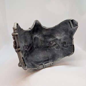 May include: A gray, textured mask with a detailed design. The mask has a weathered appearance, with visible wear and tear. The mask is likely made of a durable material and is designed to fit the face. The mask has a unique, artistic design.