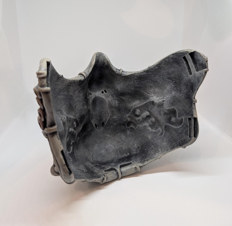 May include: A gray, textured mask with a detailed design. The mask has a weathered appearance, with visible wear and tear. The mask is likely made of a durable material and is designed to fit the face. The mask has a unique, artistic design.
