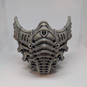 May include: A gray, intricately designed mask with a skeletal or biomechanical aesthetic. The mask features layered details, curved edges, and a symmetrical design. The color is a muted gray with subtle shading, giving it a textured appearance.