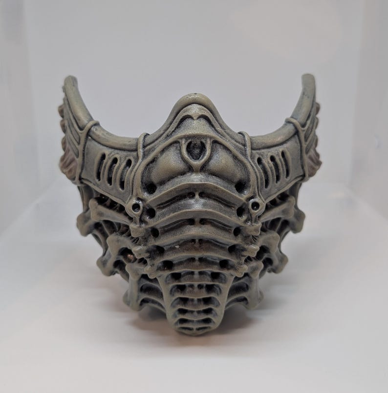 May include: A gray, intricately designed mask with a skeletal or biomechanical aesthetic. The mask features layered details, curved edges, and a symmetrical design. The color is a muted gray with subtle shading, giving it a textured appearance.