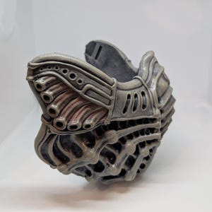 May include: A gray, intricately designed mask with a biomechanical aesthetic. The mask features multiple tubes, vents, and a textured surface, suggesting a futuristic or sci-fi theme. The color palette is primarily gray with hints of reddish-brown.