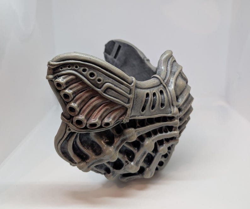 May include: A gray, intricately designed mask with a biomechanical aesthetic. The mask features multiple tubes, vents, and a textured surface, suggesting a futuristic or sci-fi theme. The color palette is primarily gray with hints of reddish-brown.
