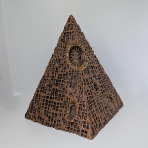 K&ouml;nnte beinhalten: Eine braune pyramidenf&ouml;rmige Skulptur mit einer strukturierten, gitterartigen Oberfl&auml;che. Die Skulptur weist kreisf&ouml;rmige, spiralf&ouml;rmige Designs auf und besteht aus einem dunkelbraunen Material. Die Pyramide ist ein dekorativer Gegenstand.