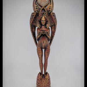 Può includere: Una statuetta umanoide color bronzo con un'estetica biomeccanica allungata. La figura poggia su una base testurizzata e presenta intricati motivi a coste su tutto il corpo, grandi strutture alari e una cresta circolare dietro la testa.