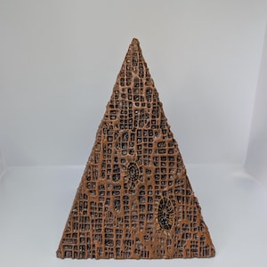 K&ouml;nnte beinhalten: Eine bronzefarbene, pyramidenf&ouml;rmige Skulptur mit einer strukturierten, gitterartigen Oberfl&auml;che. Die Skulptur weist zwei ovale, fossilartige Designs auf. Das Kunstwerk wird vor einem schlichten, wei&szlig;en Hintergrund pr&auml;sentiert.